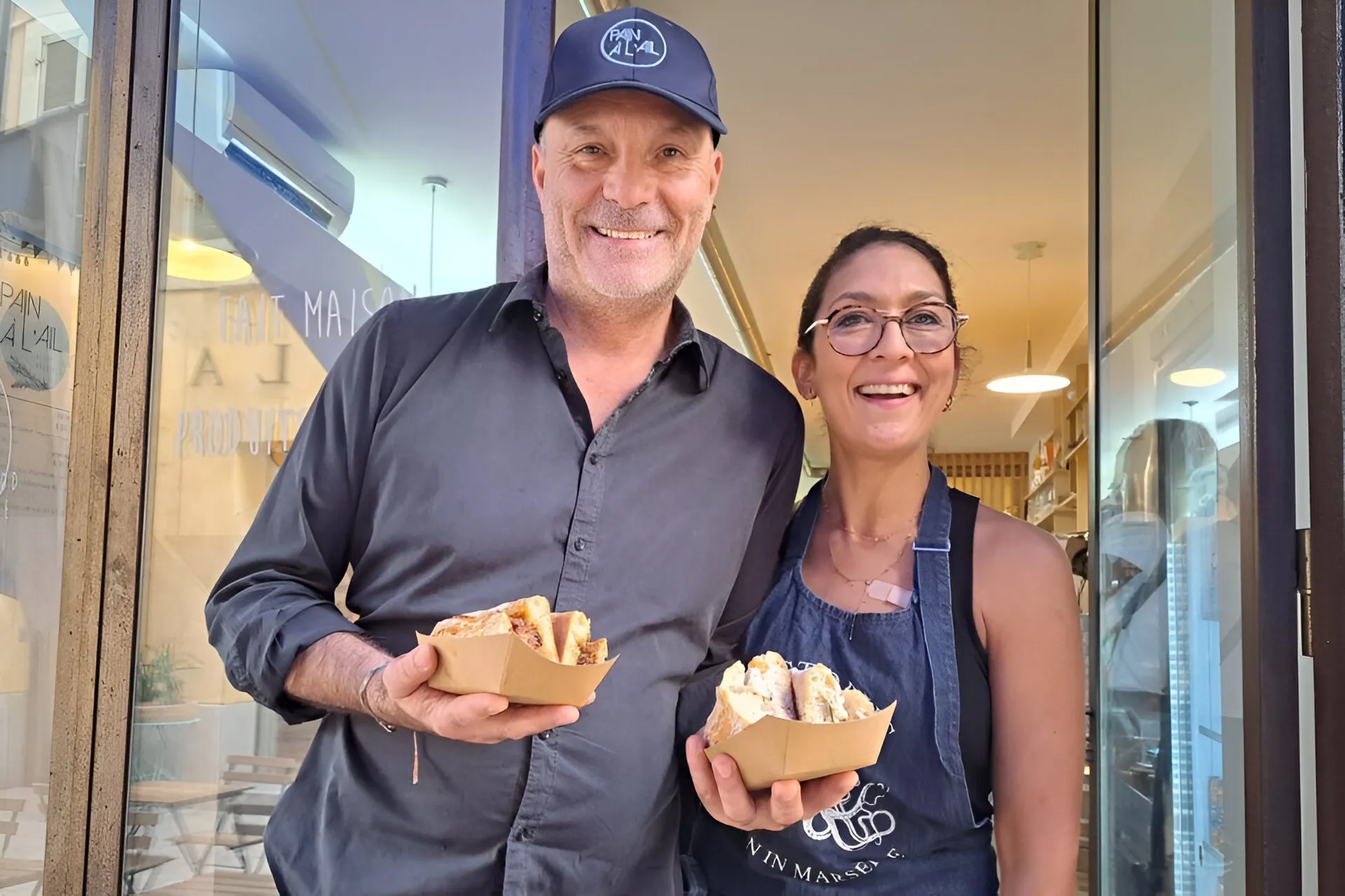 Hervé et Linda, les gérants du restaurant Pain à l’Ail, souriants devant leur établissement en tenant des plats.